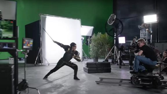 Behind the scenes of a film production with the actor playing a charging warrior in front of a green screen. There is a man operating a camera that's fixed to a track, other sound and lighting equipment is seen in the background. 
