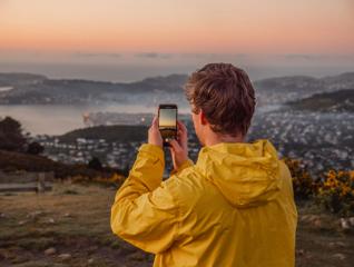 A person in a yellow windbreaker takes a picture of the sunset with their phone from Mount Kaukau.