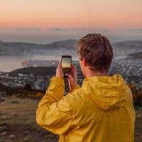 A person in a yellow windbreaker takes a picture of the sunset with their phone from Mount Kaukau.