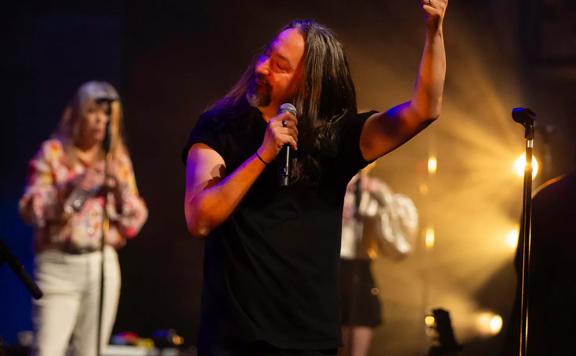 Come Together, a New Zealand-based Supergroup Tribute Band, performing on stage. The lead vocalist is holding a microphone in his right hand with his left arm raised doing a thumbs up.  