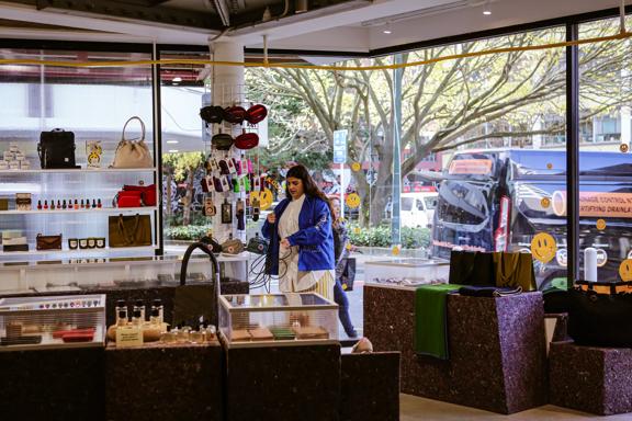 The interior of the shop Good as Gold look out on Bond Street. Glass display cases and shelves hold wallets and bags. The shop assistant stands in the background.