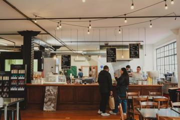 The interior of Wellington Chocolate Factory, located on Eva Street in Wellington with a large wood counter, two wood tables with four wood chairs each with three people working and two customers.