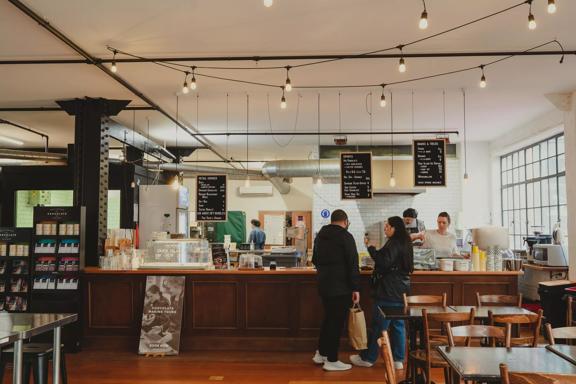 The interior of Wellington Chocolate Factory, located on Eva Street in Wellington with a large wood counter, two wood tables with four wood chairs each with three people working and two customers. 