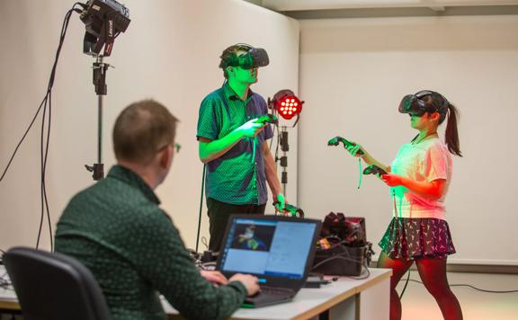 Two students wear VR headsets and use hand controls, while a lecturer watches and monitors a laptop.