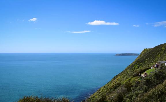 View from Boomrock lodge showing Mana island and blue water, with green bushy hills to the left and people sitting outside the lodge.