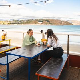 Two people sit and enjoy iced coffees and a dessert on the beach-side patio at Maranui café in Lyall Bay, Wellington.