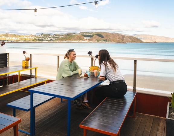 Two people sit and enjoy iced coffees and a dessert on the beach-side patio at Maranui café in Lyall Bay, Wellington.