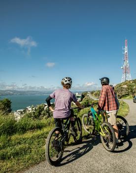 Three cyclists wearing helmets and standing next to their bikes. They are looking out into the distance across a blue stretch of water on a sunny blue-sky day.