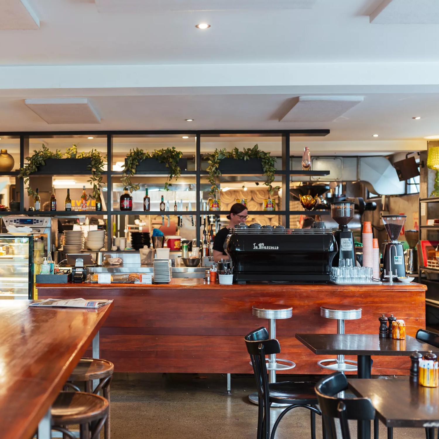 Inside Revive Café with a large wooden table and counter, with a coffee machine and glass cabinet stacked with food. Plants sit on shelving behind the counter.