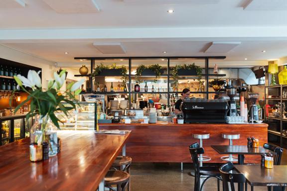 Inside Revive Café with a large wooden table and counter, with a coffee machine and glass cabinet stacked with food. Plants sit on shelving behind the counter.