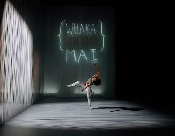 A dancer in a white outfit performs contemporary choreography under a spotlight.