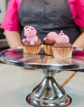 A film still from ‘Extreme Cake Sports’, a Wellington-based reality TV show that combines sports and competitive baking and involves intermediate-aged students. Three cupcakes decorated with pink pigs are on a silver desert platter.