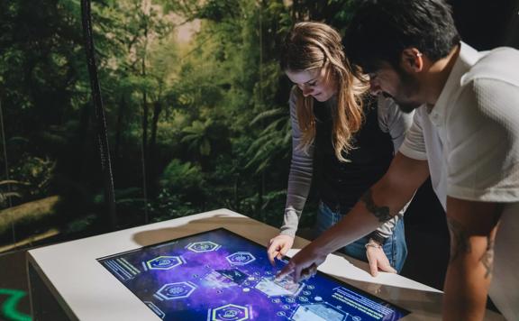 Two people looking down and pointing at an interactive display touchscreen. The wall behind them makes it look like they are surrounded by green rainforest trees.