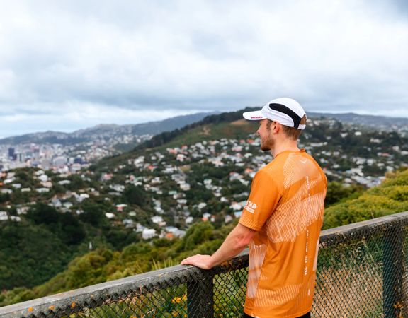 A runner wearing an orange teeshirt and white cap looks out over the hills of Wellington.