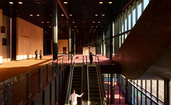 The escalators connecting the ground and first floor at the Tākina Convention Centre in Wellington. There are four people in view, brown wooden walls, black metal ceiling, floor to ceiling windows and red and dark yellow carpets. 