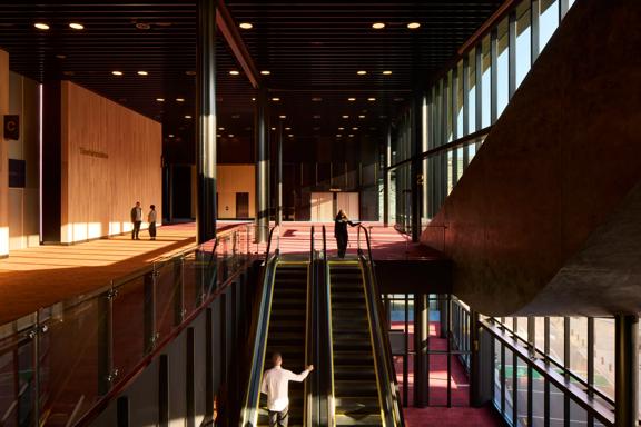 The escalators connecting the ground and first floor at the Tākina Convention Centre in Wellington. There are four people in view, brown wooden walls, black metal ceiling, floor to ceiling windows and red and dark yellow carpets. 