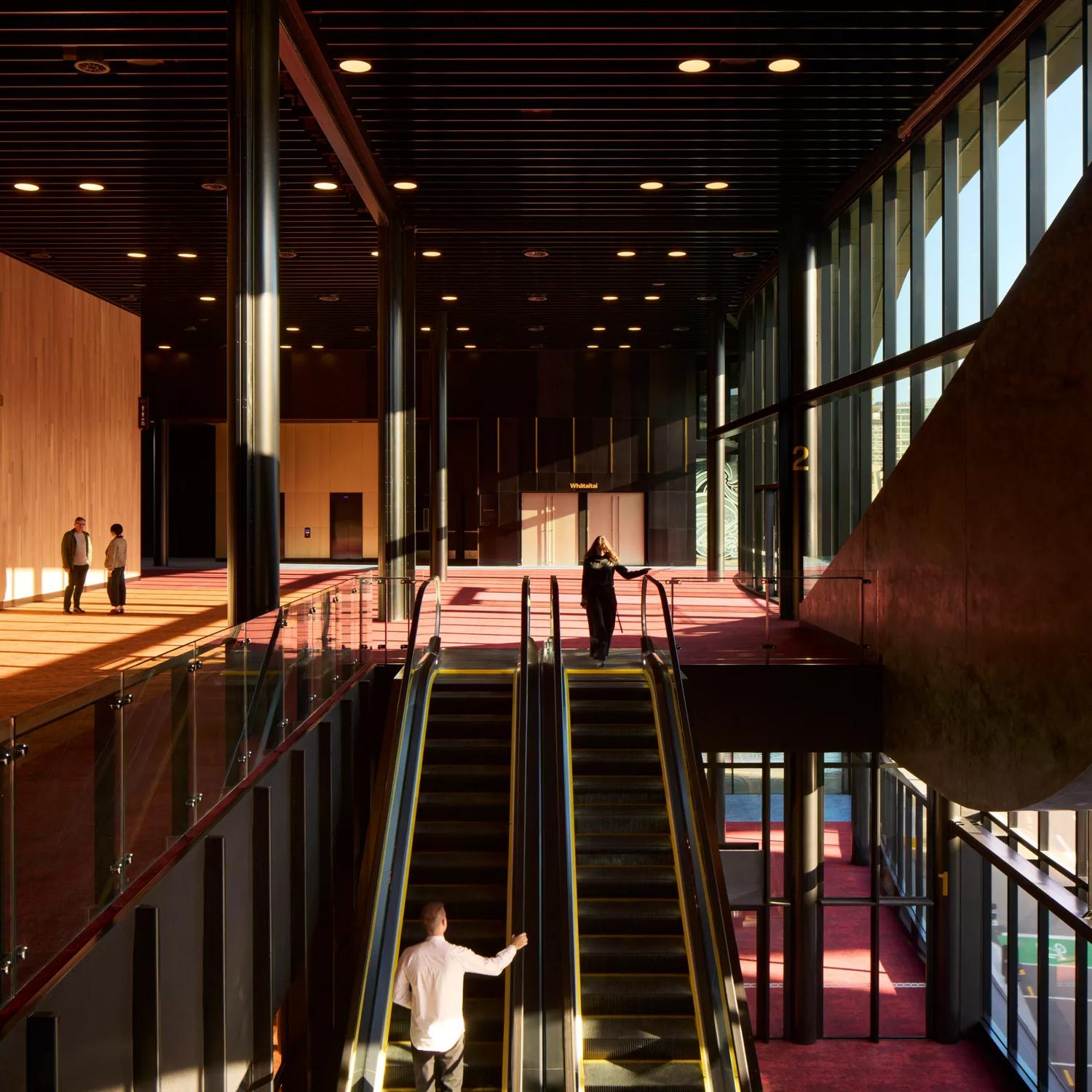 The escalators connecting the ground and first floor at the Takina Convention Centre in Wellington. There are four people in view, brown wooden walls, black metal ceiling, floor to ceiling windows and red and dark yellow carpets. 