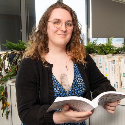 Portrait of Luna Wilkes, an inter at Cogo. They wear glasses, a black cardigan over a blue dress, and have shoulder-length wavy brown hair.