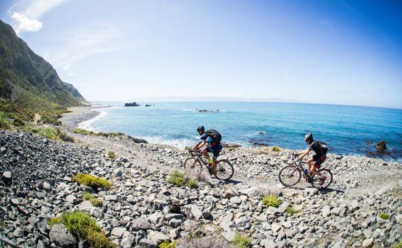 Two cyclists ride along a rocky path by Palliser Bay on the Wild Coast Trail Section in the Remutaka Cycle Trail under a sunny blue sky.