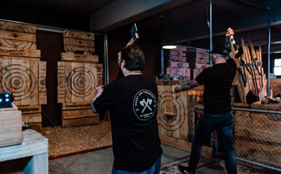 Two people side-by-side prepare to throw axes at a wooden target.