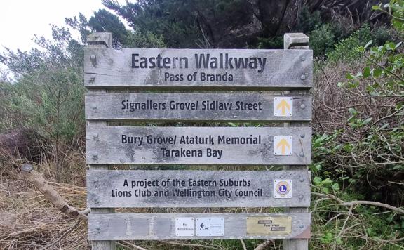 Wooden sign that reads: Eatern Walkway Pass of Branda. It has directional arrows for Signallers Grove / Sidlaw Street, Bury Grove / 
 Ataturk Memorial Tarakena Bay. A project of the Eastern Suburbs Lions Club and Wellington City Council.