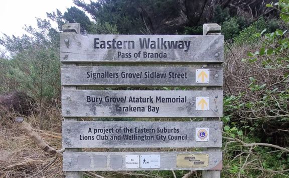 Wooden sign that reads: Eatern Walkway Pass of Branda. It has directional arrows for Signallers Grove / Sidlaw Street, Bury Grove / 
 Ataturk Memorial Tarakena Bay. A project of the Eastern Suburbs Lions Club and Wellington City Council.