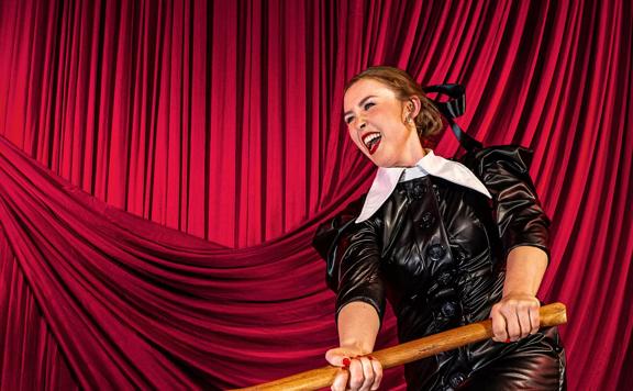 An actress portrays Lizzie Borden from 'Lizzie the Musical' posing in costume, swinging an axe in front of a red velvet curtain.