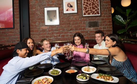 Six friends sit in a booth with sharing plates on the table. They raise their wine glasses to toast.