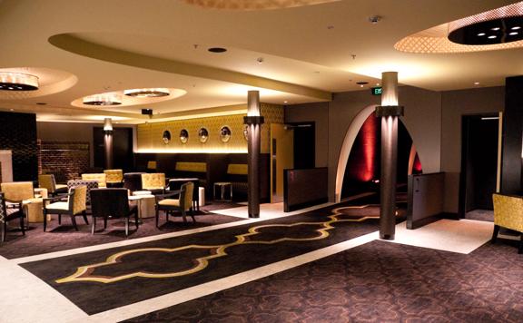 A lounge space with brown carpets an arch leading to a hallway and a seating area with small tables and armchairs.