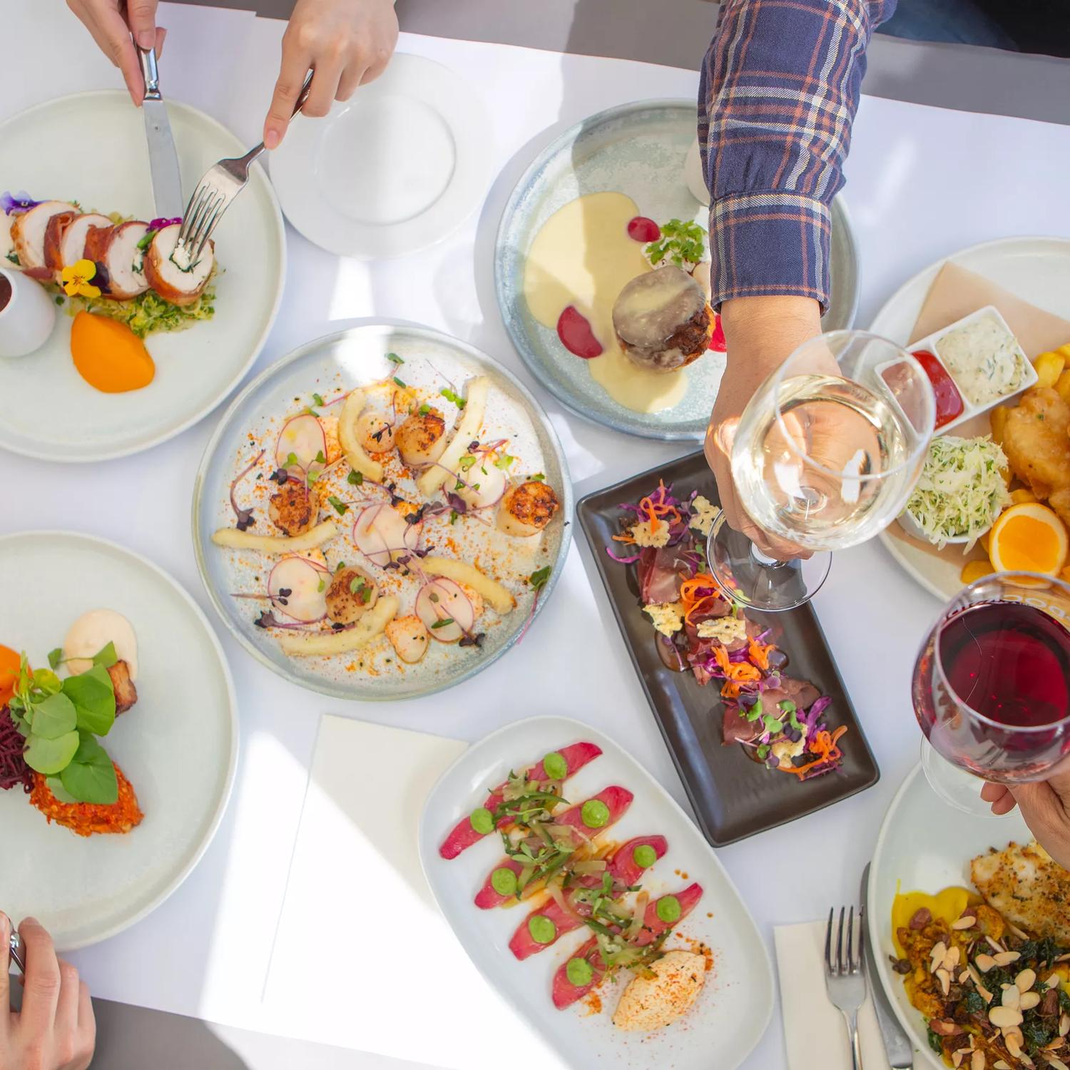 An overhead view of a delicious spread of dishes on a table at Dockside Restaurant & Bar. Four diners reach into the frame to take bites and toast their wine glasses.