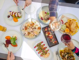 An overhead view of a delicious spread of dishes on a table at Dockside Restaurant & Bar. Four diners reach into the frame to take bites and toast their wine glasses.