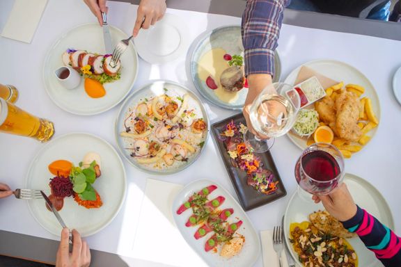 An overhead view of a delicious spread of dishes on a table at Dockside Restaurant & Bar. Four diners reach into the frame to take bites and toast their wine glasses.