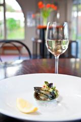 A paua shell sits on a bed of ice. In it is paua and microgreens. A lemon wedge is to the side. A glass of wine is in the background.