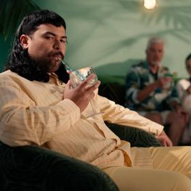A person sitting in a green velvet armchair, wearing a yellow pinstripe shirt and yellow pants, drinking a cocktail from a straw. Blurred people sit in the background mingling.