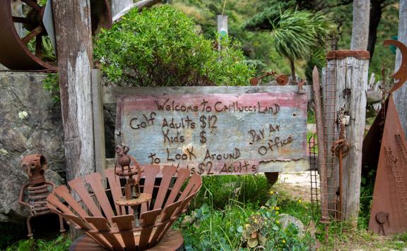 The sign at Carlucci Land, which is created from metal, says “Welcome to Carlucci Land” and lists prices. It is surrounded by metal art and plants.