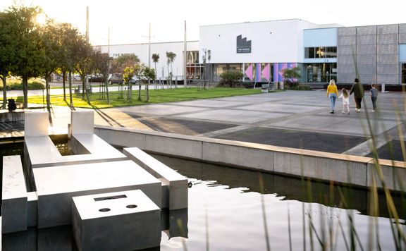 Family walking towards thre Dowse Museum. There is a large statue of a square man floating in the water.