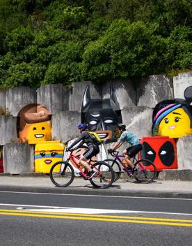 Two people on bikes passing lego figurines painted on a concrete wall on the Wainuiomata Connector Ride.