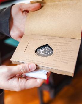 Two hands open the outer cover of a be happy chocolate bar. The packaging is brown cardstock with a tear dop shape cut out of the middle. 