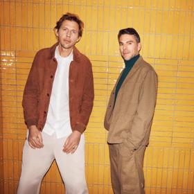 Groove Armada duo Tom Findlay and Andy Cato stand in front of an orange subway tiled wall.