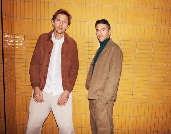 Groove Armada duo Tom Findlay and Andy Cato stand in front of an orange subway tiled wall.