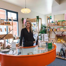 Munch Cupboard founder Anna Bordignon is dressed in a black button-down top and pants and stands behind a half-circle orange counter with a glass top in the Munch Cupboard store.