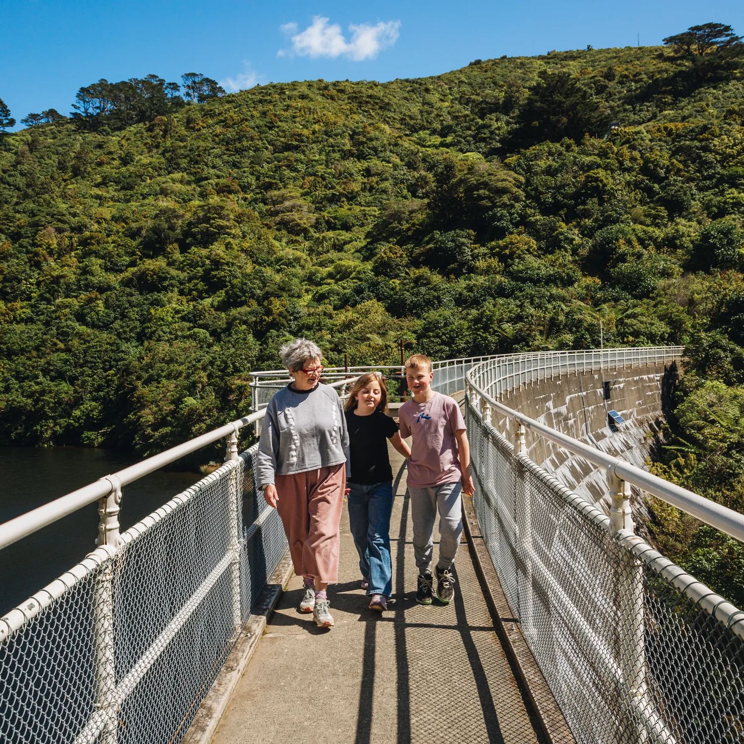 An adult and two kids walk along the top of a fenced damn at Zealandia on a sunny day.