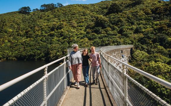 An adult and two kids walk along the top of a fenced damn at Zealandia on a sunny day.