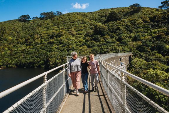 An adult and two kids walk along the top of a fenced damn at Zealandia on a sunny day.