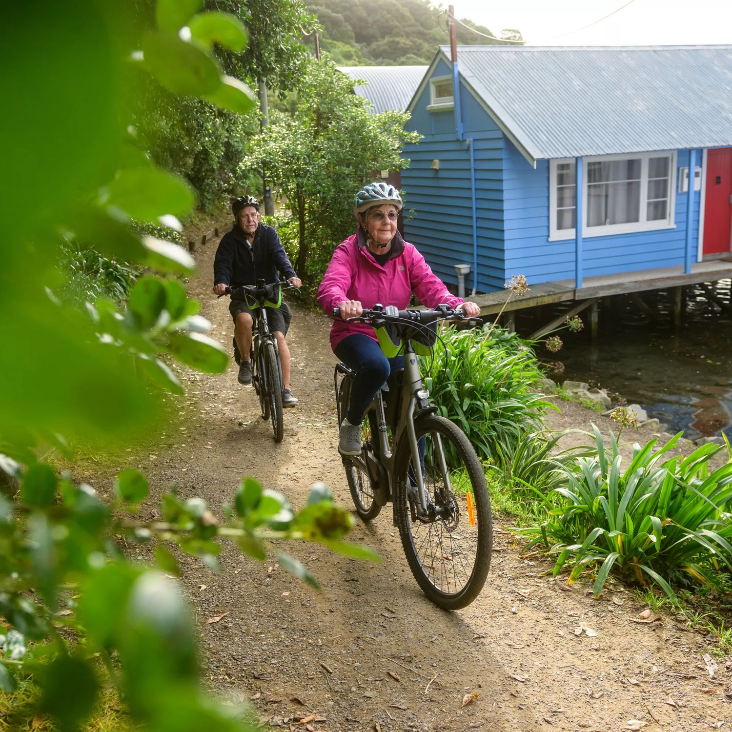 A couple on bikes cycling through a narrow path next to the boatsheds on the Camborne Walkway.