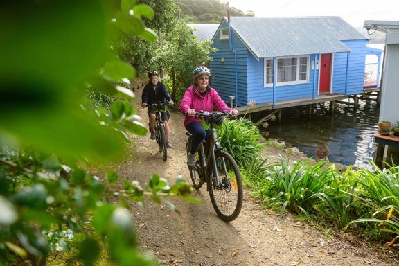 A couple on bikes cycling through a narrow path next to the boatsheds on the Camborne Walkway.