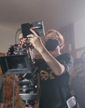 A camera operator is standing behind a industry-standard camera that's setup on a tripod. They are wearing eye glasses, a black face mask, a black tee shirt and blue jeans. 