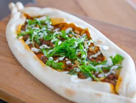 A Turkish pide with rocket and dressing drizzled on top sitting on a wooden board.