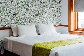 A queen-sized bed with white sheets and an olive green throw blanket draped over the foot of the bed inside a sunny room with floral wall paper.