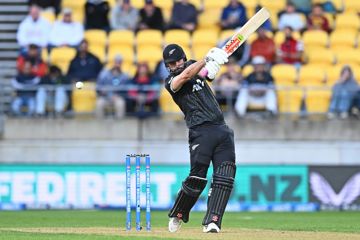 A cricket player swings a bat during a match, with stadium seating and spectators visible in the background.